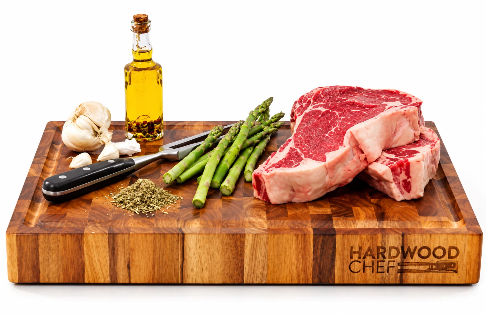 Acacia end grain board with two raw ribeye steaks, asparagus, garlic, dried herbs, a two-pronged meat fork and a corked bottle of olive oil